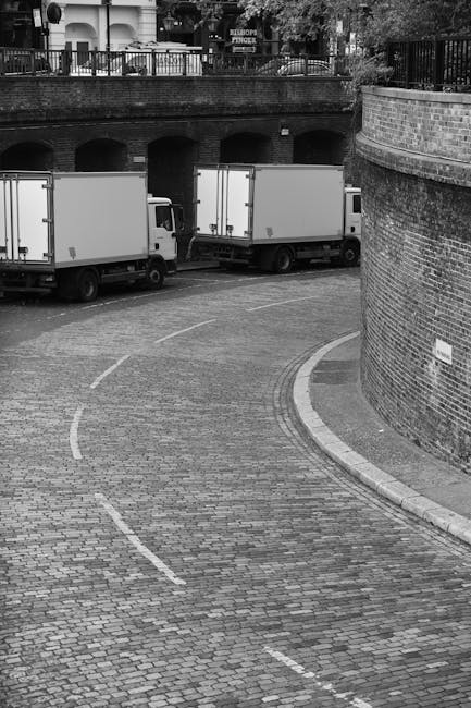 Black and white photograph depicting a curved cobblestone driveway at the entrance of an urban property. Two white box trucks are parked side by side along the left side of the driveway, positioned parallel to a brick wall that curves alongside the road. The trucks are in a stationary position, likely during a home relocation or moving process, with visible rear doors. In the background, there is a brick bridge or arched overpass with metal railings on top, above which trees and residential buildings are visible. The scene is illuminated by natural daylight, highlighting the textures of the cobblestones, brick walls, and moving vehicles. Man With a Van Deptford's professional removals service is supported by the visual context of moving logistics and furniture transport occurring at this location.