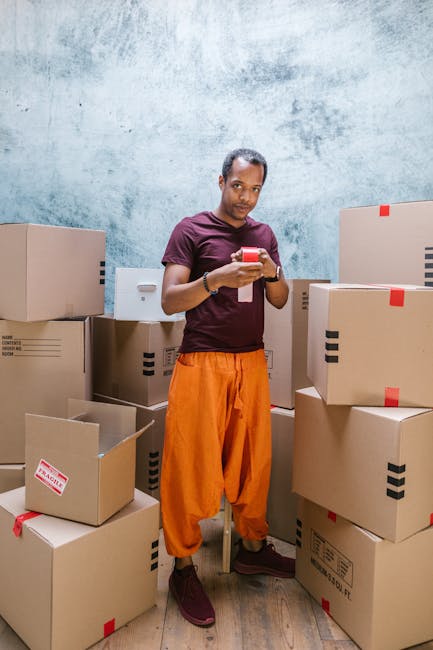 A man standing inside a room surrounded by numerous cardboard boxes of various sizes, some sealed with red tape and others open. He is wearing an orange pair of trousers, a dark purple t-shirt, and maroon shoes, holding a smartphone in his hands and looking at the camera. The boxes are stacked on the floor and some are placed on a shelf behind him. The room has a textured, light blue-grey wall and wooden flooring. This scene depicts a packing or home relocation process, relevant to furniture transport and moving logistics, with the man possibly preparing for or coordinating a house removal service by Man With a Van Deptford to help with a nearby house move or packing and moving task.