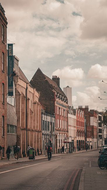 A street scene in Deptford with a row of multi-storey terraced residential buildings featuring brick facades, some with decorative window frames and chimneys, under a cloudy sky. On the pavement, ther