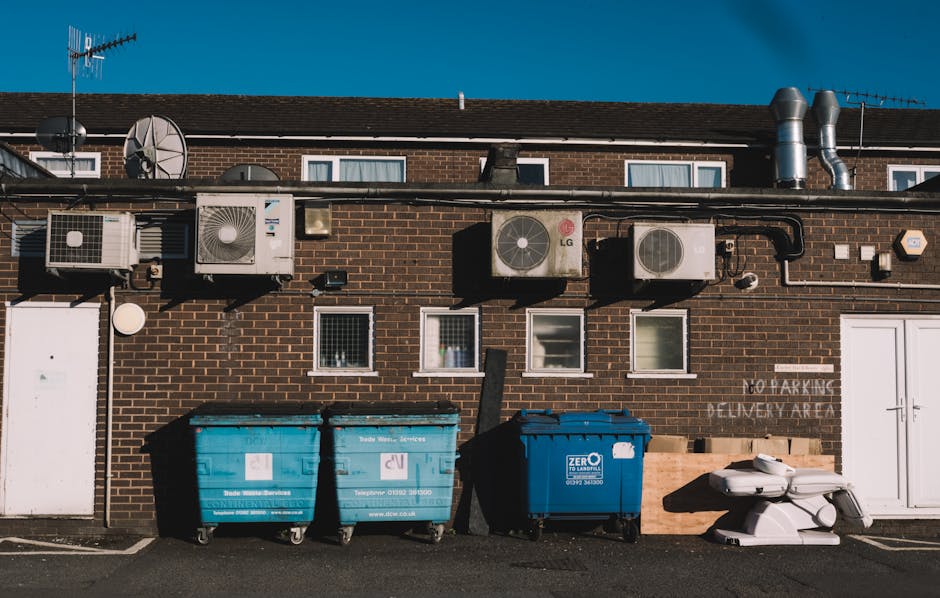 A brick building exterior showing multiple air conditioning units, satellite dishes, and ventilation pipes mounted on the upper wall, with three small windows aligned horizontally below. On the ground, there are two large blue and one black waste bins positioned side by side in front of a white door. To the right of the waste bins, a white plastic trolley with handles and wheels is placed on a paved area. The environment appears to be an outdoor yard or loading zone adjacent to the building, with clear weather under bright natural lighting. This scene illustrates a typical setup for waste disposal and loading activities related to house removals or home relocation services, as handled by Man With a Van Deptford, in accordance with local rules for skip permits and bulky waste removals as outlined on the specified webpage.