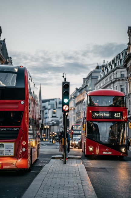 A busy street scene in Deptford during daytime featuring two red double-decker buses, one on the left and another on the right, with the bus on the right displaying the route number 453 and 'Deptford Bridge' as its destination. The buses are positioned on a paved road with a central pedestrian crossing that has tactile paving, and a traffic light in the middle displaying a green signal. Modern and historic buildings line the street, with a cloudy sky overhead creating a diffused lighting effect. The scene is typical of urban house removal environments where transport and logistics are involved in home relocation services, supported by companies like Man With a Van Deptford, which handles furniture transport, packing, loading, and moving processes within the area.