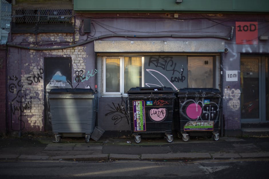 A brick building exterior showing multiple air conditioning units, satellite dishes, and ventilation pipes mounted on the upper wall, with three small windows aligned horizontally below. On the ground, there are two large blue and one black waste bins positioned side by side in front of a white door. To the right of the waste bins, a white plastic trolley with handles and wheels is placed on a paved area. The environment appears to be an outdoor yard or loading zone adjacent to the building, with clear weather under bright natural lighting. This scene illustrates a typical setup for waste disposal and loading activities related to house removals or home relocation services, as handled by Man With a Van Deptford, in accordance with local rules for skip permits and bulky waste removals as outlined on the specified webpage.