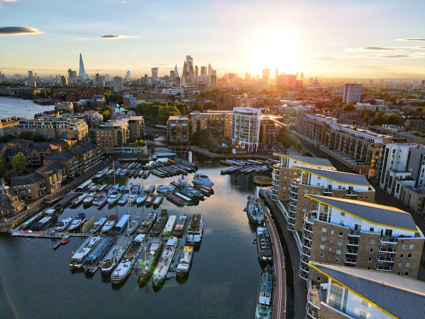 Aerial view of a marina in an urban riverside area during sunset, showing numerous boats moored along the docks, with modern residential apartment buildings lining the water's edge. The city skyline, including high-rise buildings and recognizable skyscrapers such as The Shard, is visible in the background under a sky with scattered clouds and warm, golden sunlight. The scene captures the busy environment of riverside homes, suitable for house removals or furniture transport, with the reflection of the buildings and sky on the calm water. This image illustrates the proximity to the river, typical of riverside property locations, and the urban setting where home relocation services might be undertaken, as provided by companies like Man With a Van Deptford.