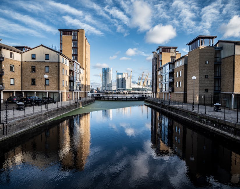 A river with calm, reflective water runs between modern, multi-story residential buildings constructed with light brown brick and concrete, positioned along the canal's edges. The buildings feature small balconies and numerous windows facing the water, with a few cars parked along the promenade and black metal fencing lining the waterway. In the background, a bridge spans the river, and beyond that, contemporary high-rise buildings and cranes are visible against a partly cloudy sky, indicating an urban riverside setting in Deptford. The scene is well-lit with natural daylight, highlighting the architecture and the tranquil water surface, which mirrors the buildings, sky, and clouds. This image evokes a scene of home relocation and furniture transport preparations, typical of a house removals service operating in the Deptford Creekside area, with a focus on moving logistics dependably handled by companies like Man With a Van Deptford.