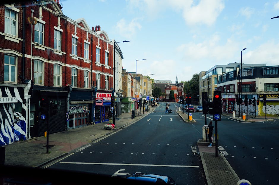 A daytime street scene in Deptford, SE8, showing a mix of red brick and modern buildings with retail shops and cafes lining the pavement. In the foreground, a section of a black paint-streaked vehicle is visible, indicating the photo was taken from inside a moving van used for house removals. On the right side, a pedestrian waits at a traffic light, while several cardboard boxes and wrapped furniture are placed along the sidewalk, suggesting packing or home relocation activities. There are traffic signals and street markings, including bus and cycle lanes, with parked cars in the distance. The sky is partly cloudy with natural daylight illuminating the urban environment. This scene reflects the process of furniture transport and loading during a professional move, highlighting elements typical of house removals in the area. Man With a Van Deptford occasionally appears to support local house removals and transport logistics.