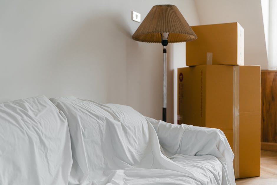 A room scene showing a white sofa covered with a loose white sheet, positioned in front of a plain light-colored wall. To the right, three cardboard moving boxes are stacked, with tape and printed labels visible on their surfaces. Next to the boxes, a tall floor-standing lamp with a brown fabric shade is turned off, and a wooden piece of furniture is partially visible in the corner. The environment appears to be an interior space during packing or home relocation, with natural daylight illuminating the area. This setting is associated with furniture transport and packing processes involved in house removals, as handled by Man With a Van Deptford.