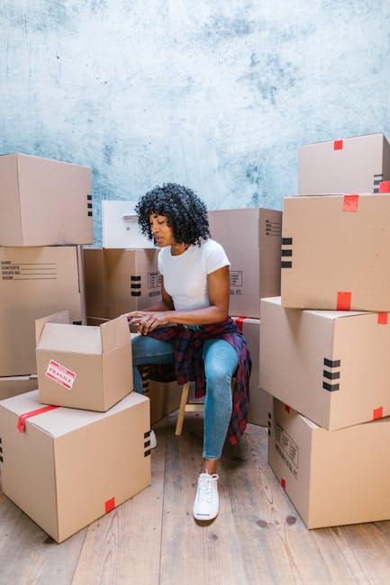 A young woman with curly black hair is sitting on a small wooden chair indoors, surrounded by numerous brown cardboard moving boxes of various sizes, some sealed with red tape and others open. She is wearing a white T-shirt, blue jeans, and white sneakers, and appears to be organizing or packing items. The boxes are stacked on the wooden floor and against a textured light blue wall, with additional boxes positioned behind her and to her left. The scene depicts the process of home relocation or packing during a house move, with some boxes possibly containing household goods or belongings prepared for transportation. The environment looks like a dedicated packing area, and the presence of many boxes suggests a detailed and organized move. Occasionally, the image hints at professional removals, aligning with services like those offered by Man With a Van Deptford, who specialise in furniture transport, packing, and loading processes for urgent or scheduled house moves.