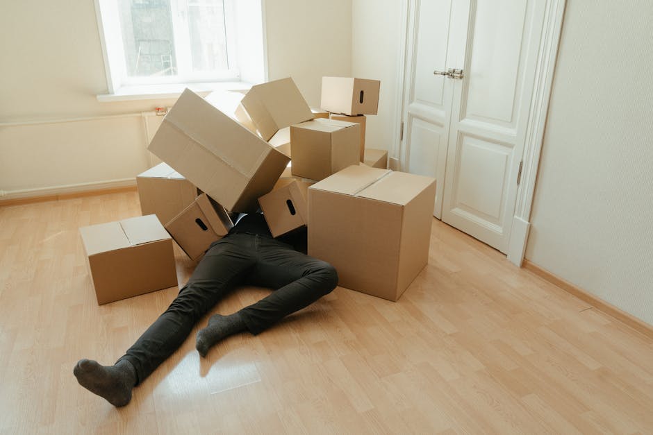 A room scene showing a white sofa covered with a loose white sheet, positioned in front of a plain light-colored wall. To the right, three cardboard moving boxes are stacked, with tape and printed labels visible on their surfaces. Next to the boxes, a tall floor-standing lamp with a brown fabric shade is turned off, and a wooden piece of furniture is partially visible in the corner. The environment appears to be an interior space during packing or home relocation, with natural daylight illuminating the area. This setting is associated with furniture transport and packing processes involved in house removals, as handled by Man With a Van Deptford.