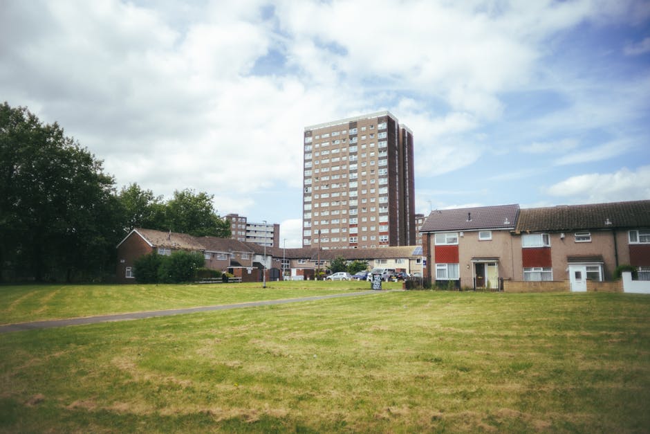 A wide outdoor view of a residential area in Deptford, showing a large grassy open space with a paved footpath running through it. In the background, there are several single-story houses with brick and stucco exteriors, some with red and white window shutters, and a multi-storey modern apartment building with numerous windows. To the left, a cluster of leafy trees adds greenery to the scene. The sky is partly cloudy with patches of blue visible. This setting illustrates a typical neighborhood environment where house removals and furniture transport might take place, with the open area potentially used for loading and unloading goods during a home relocation, as demonstrated by the presence of parked cars and a moving company such as Man With a Van Deptford in the vicinity.