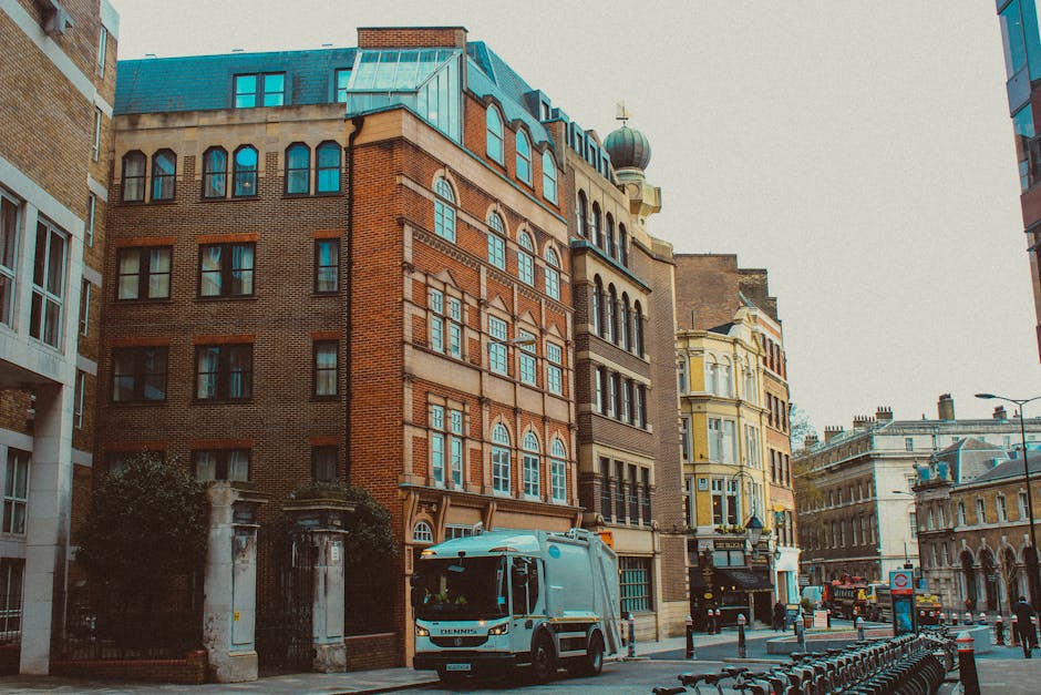 A street scene in Deptford showing several multi-storey buildings with brick and ornate facades, some with bay windows and decorative architectural elements. In the foreground, a light blue van from Man With a Van Deptford is parked on the pavement near the entrance of a property, with a few cardboard boxes and packed items visible inside. A few wrapped furniture pieces and large cardboard cartons are being loaded into or unloaded from the van, with a person’s hands visible gripping a strap to secure the items during the home relocation process. The street is lined with metal bollards and bicycles parked along the curb, while other vehicles and buildings are visible in the background under natural daylight. The scene reflects typical elements involved in packing, furniture transport, and moving logistics associated with house removals in the Lewisham area, following council guidelines for organized and efficient home moves.