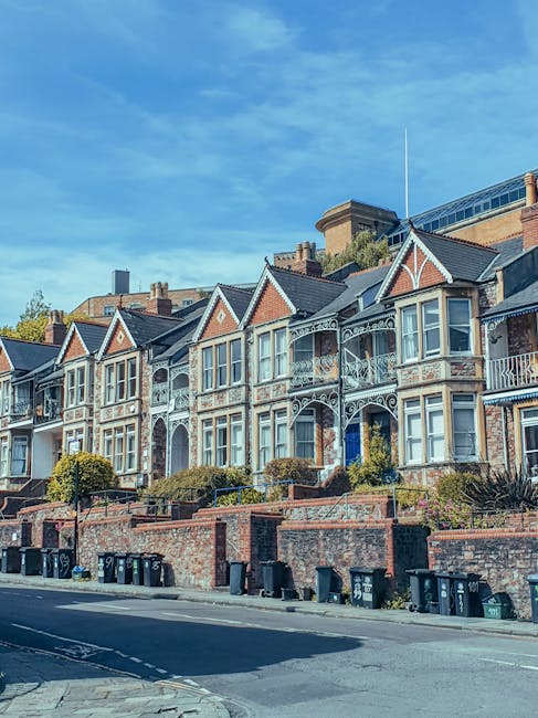 A wide outdoor view of a residential area in Deptford, showing a large grassy open space with a paved footpath running through it. In the background, there are several single-story houses with brick and stucco exteriors, some with red and white window shutters, and a multi-storey modern apartment building with numerous windows. To the left, a cluster of leafy trees adds greenery to the scene. The sky is partly cloudy with patches of blue visible. This setting illustrates a typical neighborhood environment where house removals and furniture transport might take place, with the open area potentially used for loading and unloading goods during a home relocation, as demonstrated by the presence of parked cars and a moving company such as Man With a Van Deptford in the vicinity.