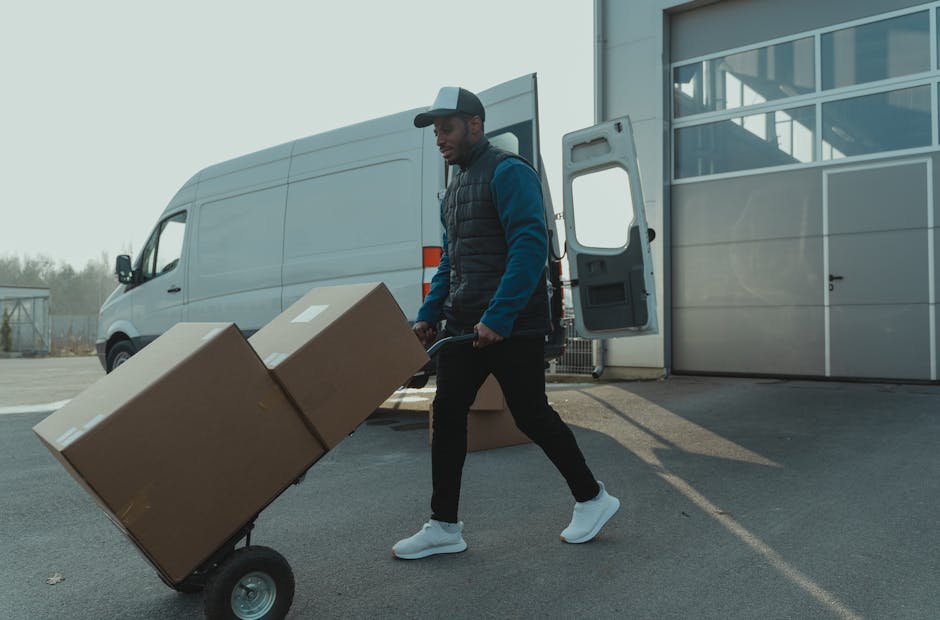 A man wearing a baseball cap, a dark blue jacket with a padded vest, and white sneakers is shown lifting a large cardboard box onto a hand truck outside a white cargo van, which is parked near a modern commercial building with large glass windows. The open rear doors of the van reveal additional boxed items and packing materials inside. The scene takes place on an outdoor paved area during daylight, with clear weather and distant trees visible in the background. The man appears engaged in a home relocation or furniture transport activity, with the logistics process of packing and loading moving boxes as part of a professional removals service, such as that provided by Man With a Van Deptford.