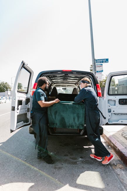 Two movers from Man With a Van Deptford are seen loading a large piece of furniture, covered with a dark green fabric blanket, into the back of a black van parked on a street. The van's rear doors are open, revealing the interior designated for furniture transport, with some packaging materials visible inside. Both workers are dressed in dark uniforms; one wears glasses and a cap, while the other has short dark hair. They are positioned on either side of the item, carefully carrying it into the vehicle, during a home relocation or furniture transport process on a clear day. The scene takes place outdoors on a pavement, with a street sign and other parked vehicles visible in the background. This image illustrates the loading process during a professional house removal service, consistent with the offerings of Man With a Van Deptford for same-day removals, supporting the logistical steps involved in packing and moving furniture during a house move.