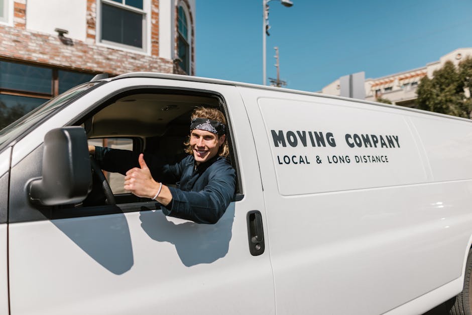 A man wearing a baseball cap, a dark blue jacket with a padded vest, and white sneakers is shown lifting a large cardboard box onto a hand truck outside a white cargo van, which is parked near a modern commercial building with large glass windows. The open rear doors of the van reveal additional boxed items and packing materials inside. The scene takes place on an outdoor paved area during daylight, with clear weather and distant trees visible in the background. The man appears engaged in a home relocation or furniture transport activity, with the logistics process of packing and loading moving boxes as part of a professional removals service, such as that provided by Man With a Van Deptford.