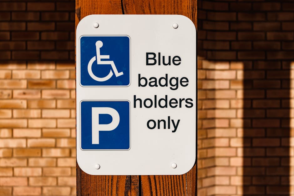 A rectangular white sign mounted on a wooden post that displays a blue accessibility symbol for wheelchair users at the top, followed by black text that reads 'Blue badge holders only' and a blue parking symbol with a white 'P'. The sign is situated outdoors against a background of a brick wall with a reddish-brown hue, and sunlight casting shadows on the wall and sign. The sign likely indicates designated parking spaces for blue badge holders, which may be relevant for house removals and home relocation services, such as those provided by Man With a Van Deptford, in compliance with parking and loading restrictions in Deptford.