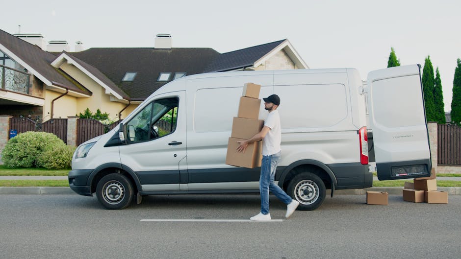 A man with a beard, dressed in a white t-shirt, jeans, and white sneakers, is loading three packed cardboard boxes into the side cargo area of a white commercial van parked on a residential street. The van is positioned parallel to the pavement, with the rear door open, revealing the interior for furniture transport and packing materials. Several additional cardboard boxes rest on the ground nearby, some stacked on top of each other. The background features modern houses with pitched roofs, a fenced garden with bushes, and tall, narrow trees, under bright daylight with clear weather. The scene depicts a home relocation or packing and moving process, with the van belonging to Man With a Van Deptford, a company providing removals and moving services.