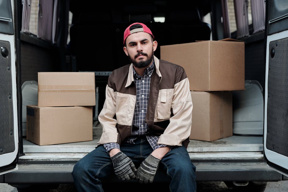 A man with a beard, wearing a red baseball cap backwards, a checkered shirt under a beige and brown jacket, and striped gloves, sitting on the edge of an open vehicle with a dark interior. Behind him are several cardboard boxes, some stacked and others placed on the floor of the van, which is parked outside a building. The scene suggests a home relocation or moving process, with packing materials visible in the form of cardboard boxes ready for furniture transport or household packing and moving services. The lighting indicates natural daylight, and the environment appears to be an outdoor loading area, consistent with house removal activities carried out by Man With a Van Deptford.