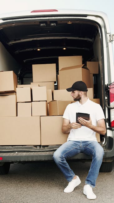 A man with a beard, dressed in a white t-shirt, jeans, and white sneakers, is loading three packed cardboard boxes into the side cargo area of a white commercial van parked on a residential street. The van is positioned parallel to the pavement, with the rear door open, revealing the interior for furniture transport and packing materials. Several additional cardboard boxes rest on the ground nearby, some stacked on top of each other. The background features modern houses with pitched roofs, a fenced garden with bushes, and tall, narrow trees, under bright daylight with clear weather. The scene depicts a home relocation or packing and moving process, with the van belonging to Man With a Van Deptford, a company providing removals and moving services.