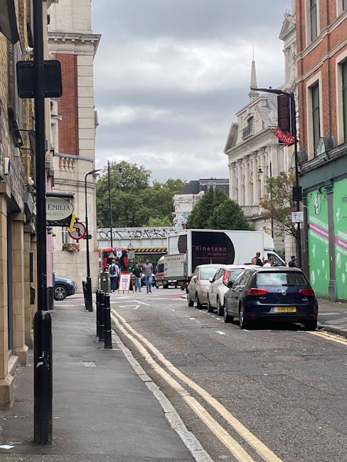 A narrow urban street scene in Deptford during daytime with parked cars along the roadside and a white moving van positioned on the carriageway, partially obscured by other vehicles. The street is lined with multi-storey buildings, including a historic white stone structure with classical architectural features, and shops with signs, one of which is green and prominent on the right side. Pedestrians are visible near a crossing area, some standing and others walking, with a few people gathered around the moving van. The pavement is marked by black bollards, and overhead traffic lights and street signage are present. The sky is overcast with grey clouds, and in the background, there are trees and additional urban buildings, indicating a lively area suitable for house relocations, furniture transport, and packing during home moves. The scene reflects a typical street setup where a removal company like Man With a Van Deptford might assist with home relocation logistics.