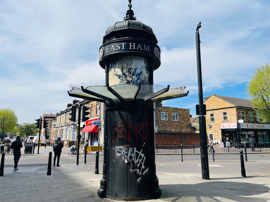 A cylindrical public notice board with a domed top displaying the words 'East Ham' is situated on a paved sidewalk at a busy street corner in daytime. The structure features a black metal frame and mesh, with visible graffiti tags on its lower section. Surrounding it are urban elements including black bollards lining the sidewalk, and a black street pole nearby. Pedestrians are walking on the pavement; some are carrying bags or making their way across the intersection. Behind the notice board, several brick buildings with shopfronts, including a nail salon and a bakery, are visible, with a mix of trees and cars in the background. The sky is partly cloudy with patches of blue, and the scene suggests a lively area consistent with typical city streets involved in home relocations or furniture transport activities, where visualising loading procedures or moving equipment outdoors is common.