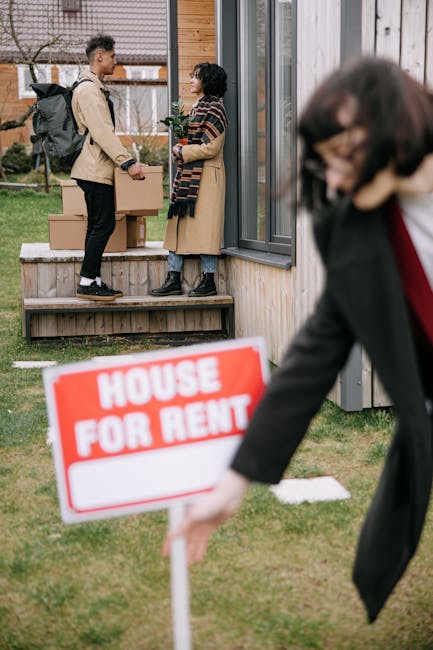 Two individuals standing on the wooden steps outside a residential property engaged in a conversation during a home relocation process. One person, carrying a cardboard box, is dressed in casual clothing and wearing a backpack, while the other, also holding a box, is wearing a coat and glasses. The scene is set near a modern building with glass doors and windows, indicating an inside-outside transition area. In the foreground, a woman dressed in a black jacket is partially visible, walking past while holding a 'House for Rent' sign with a red border and white background. The background shows a lawn with a few small trees and shrubs, and additional cardboard boxes stacked on the ground near the property. This image captures elements of packing, furniture transport, and scheduling involved in house removals, aligning with services provided by Man With a Van Deptford, as part of their house removals and moving logistics.