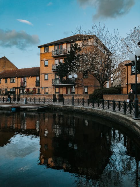 View of a multi-storey brick residential building situated alongside a waterway, with reflections visible in the calm water. The building features several balconies with metal railings and large windows, surrounded by trees and street lamps on a paved walkway. Some individuals are walking along the pathway, with one person wearing a dark coat visible near the right edge of the scene. The sky is partly cloudy with patches of blue, indicating a clear day. This image captures an urban riverside setting potentially involved in home relocation activities, with an emphasis on the surrounding environment in Surrey Canal estate, relevant to house removals services by Man With a Van Deptford.