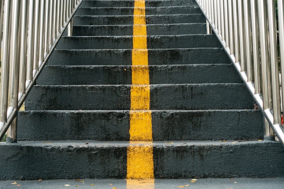 Close-up view of a concrete outdoor staircase with eight steps, painted in black with a single yellow safety line running down the center. The staircase is flanked by shiny metal handrails on both sides. The steps appear slightly wet with some small leaves scattered across them, indicating recent rain or damp conditions. The image is taken from the bottom looking upward, emphasizing the incline and the central yellow marking. This type of stairs may pose access challenges during a home relocation or furniture transport due to their steepness and narrow width, and careful handling or alternative access solutions could be required. Such stairs are common in residential or apartment building exteriors, requiring professional removals services like Man With a Van Deptford to ensure safe loading and unloading of furniture or boxes during packing and moving processes. The environment suggests an outdoor setting, possibly in an urban area, with partial greenery visible at the top edges of the image. Lighting appears natural, with daylight creating a clear, well-lit scene.