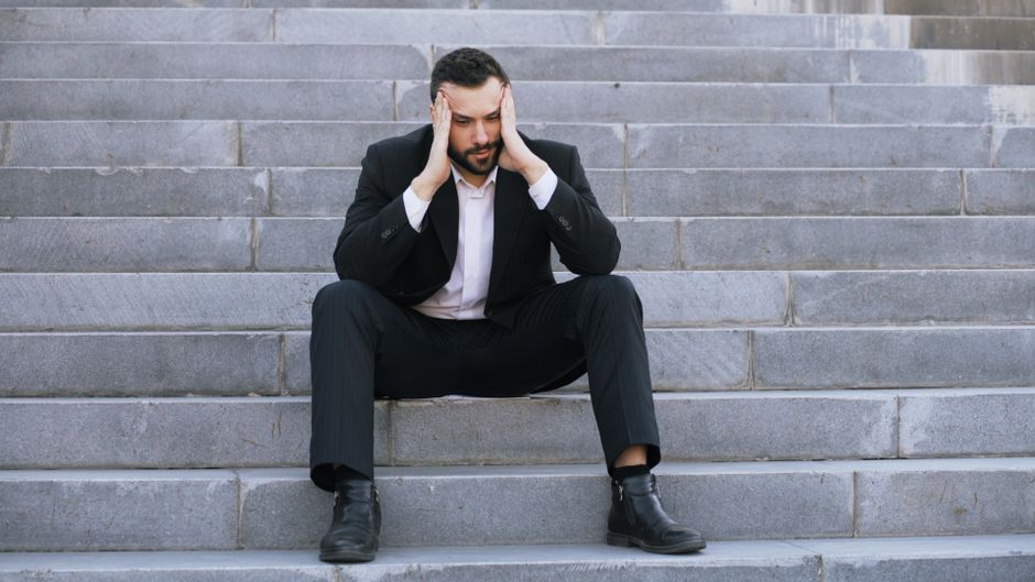 A man dressed in a black suit with a white shirt is sitting on a set of outdoor stone stairs, appearing stressed with his hands on his head. The stairs are made of grey stone, with a consistent pattern of rectangular steps that lead upward and out of view. The man’s facial expression shows concern, and he is positioned centrally in the image, with his legs spread apart and feet flat on the ground. The setting appears to be outside a property, possibly indicating a home or office building, which could pose challenges during furniture transport or home relocation processes. This scene is relevant for discussing house removals, particularly when facing stairs access problems, and highlights the importance of professional removal services like those provided by Man With a Van Deptford. The overall environment is well-lit with natural daylight, emphasizing the steps' texture and the man's attire, and suggesting the need for careful handling of furniture and packing materials when navigating staircases during a moving project.