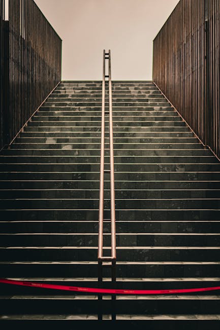 Close-up view of a concrete outdoor staircase with eight steps, painted in black with a single yellow safety line running down the center. The staircase is flanked by shiny metal handrails on both sides. The steps appear slightly wet with some small leaves scattered across them, indicating recent rain or damp conditions. The image is taken from the bottom looking upward, emphasizing the incline and the central yellow marking. This type of stairs may pose access challenges during a home relocation or furniture transport due to their steepness and narrow width, and careful handling or alternative access solutions could be required. Such stairs are common in residential or apartment building exteriors, requiring professional removals services like Man With a Van Deptford to ensure safe loading and unloading of furniture or boxes during packing and moving processes. The environment suggests an outdoor setting, possibly in an urban area, with partial greenery visible at the top edges of the image. Lighting appears natural, with daylight creating a clear, well-lit scene.